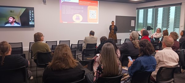 A person speaks in front of a lecture room of people seated and watching. On a screen we see an online speaker and community also joining.
