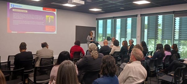 a person speaks at the front of a seminar room that is full of people looking at the speaker