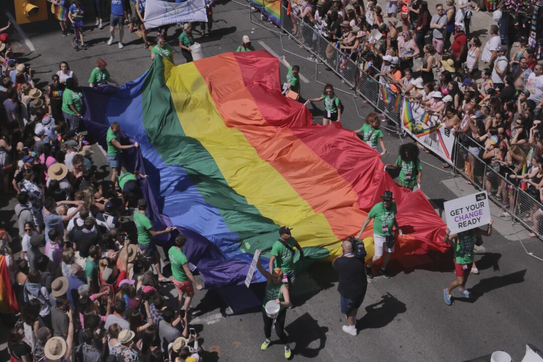 people celebrate and protest at a pride parade, there is a large rainbow flag being held up by attendees